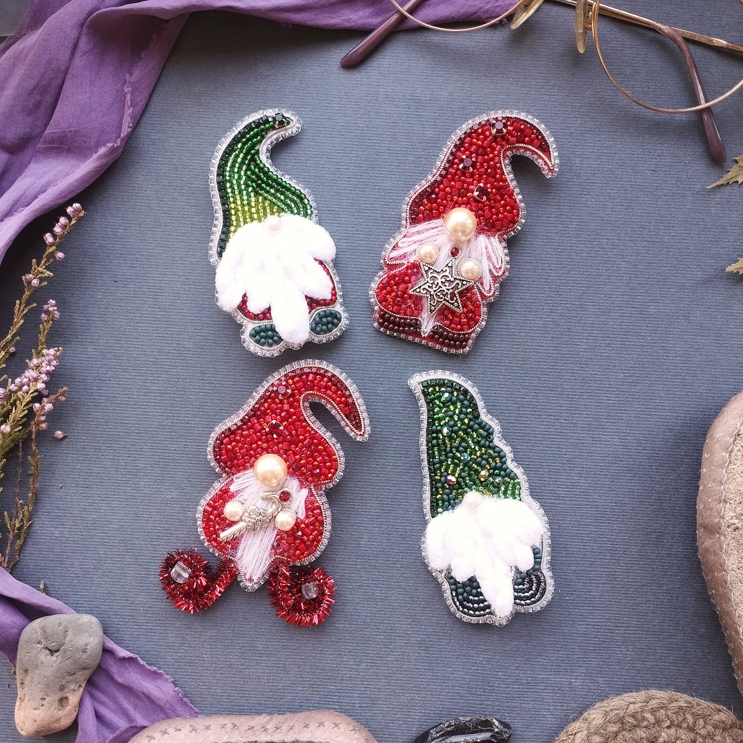 four ornate, beaded or embroidered gnome-shaped brooches or pins arranged on a gray surface.