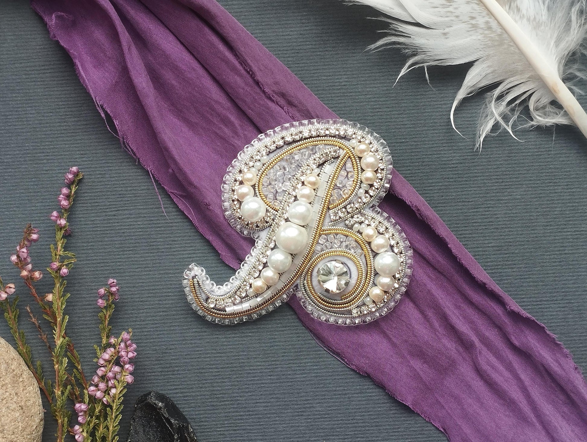 a decorative monogrammed letter "B" adorned with pearls and rhinestones, placed on a purple fabric.