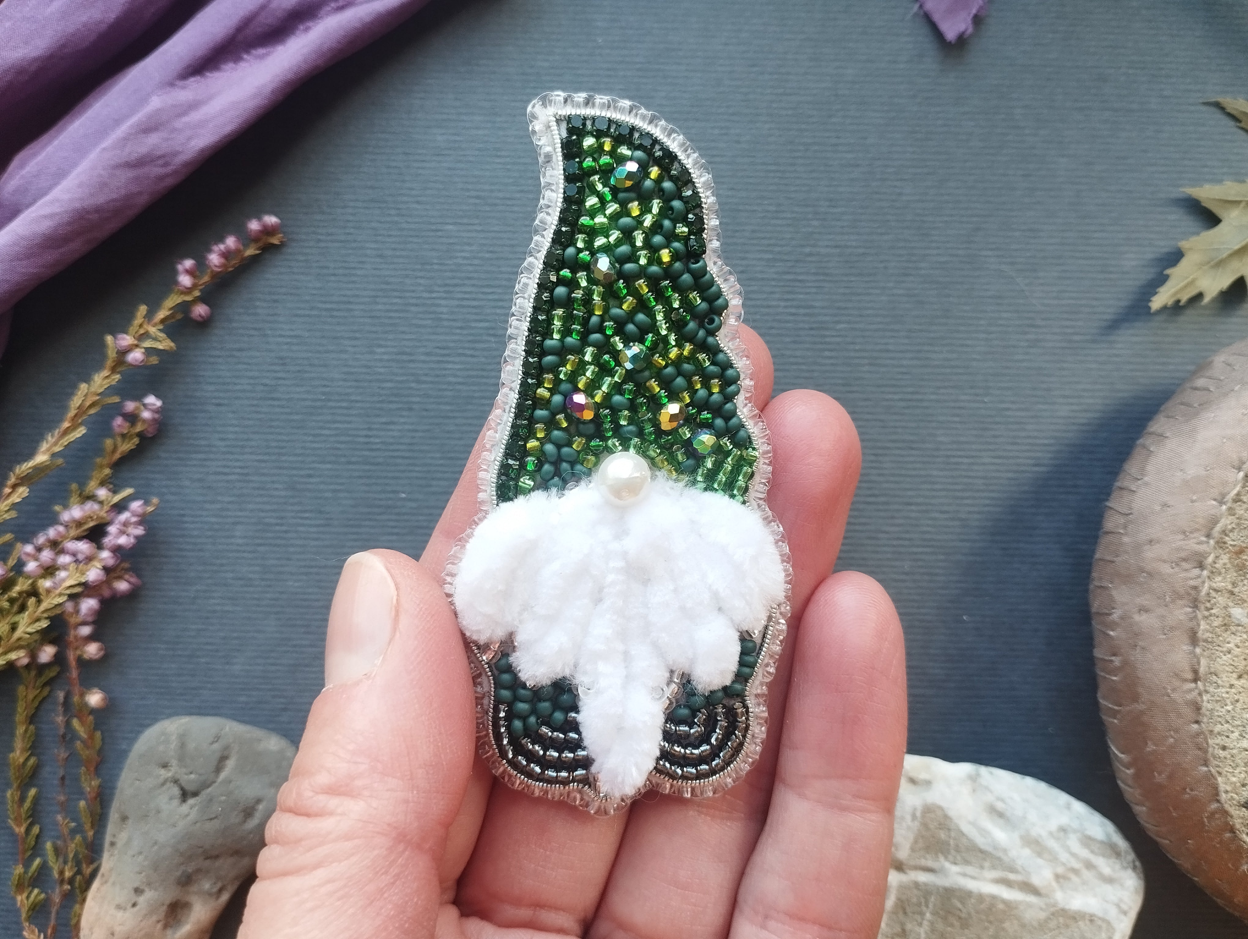 A hand holding a small, intricately designed ornament made of green and white beads, with a white flower-like design on the front.