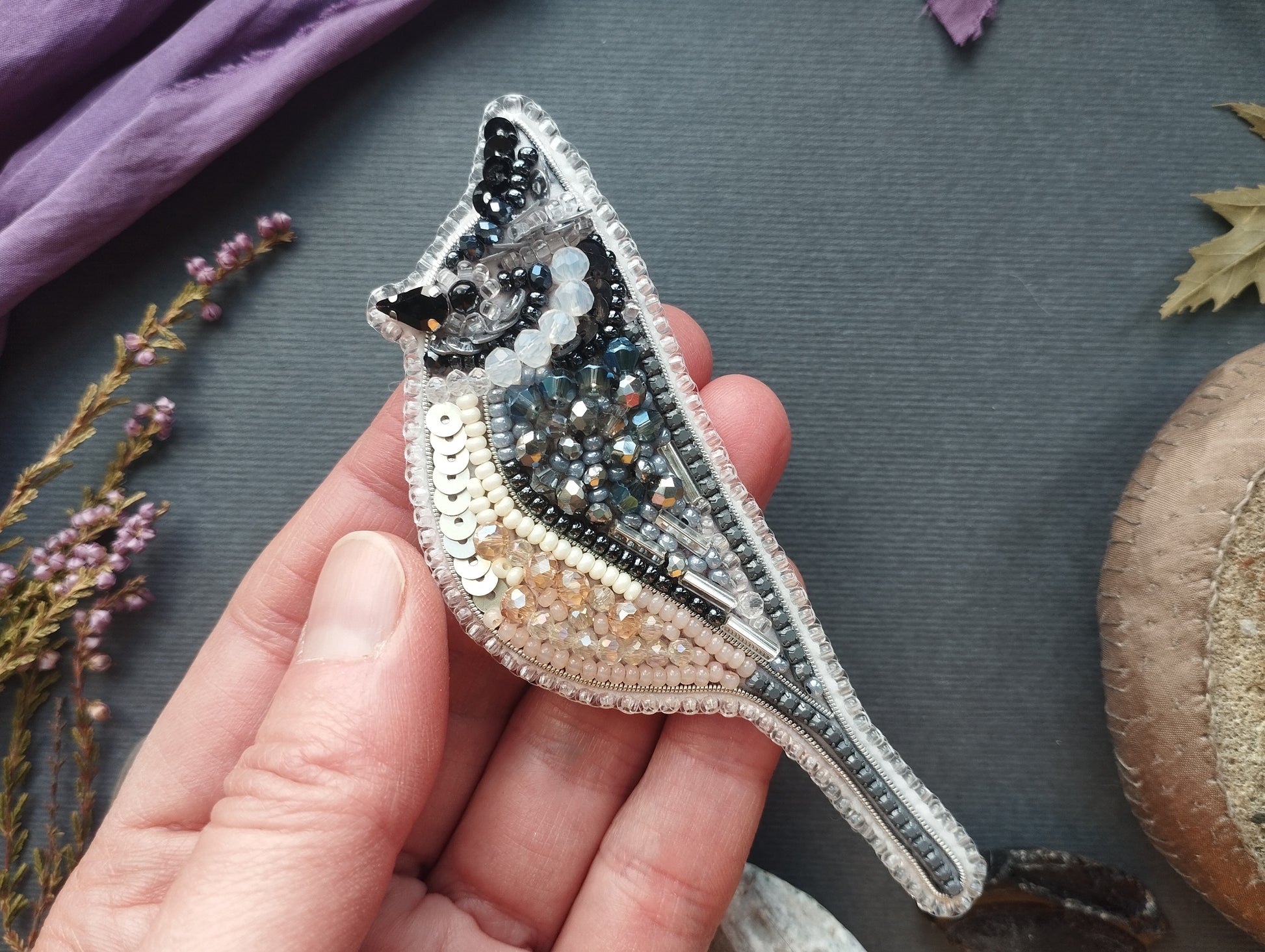 A hand is holding a decorative beaded bird brooch against a dark background.