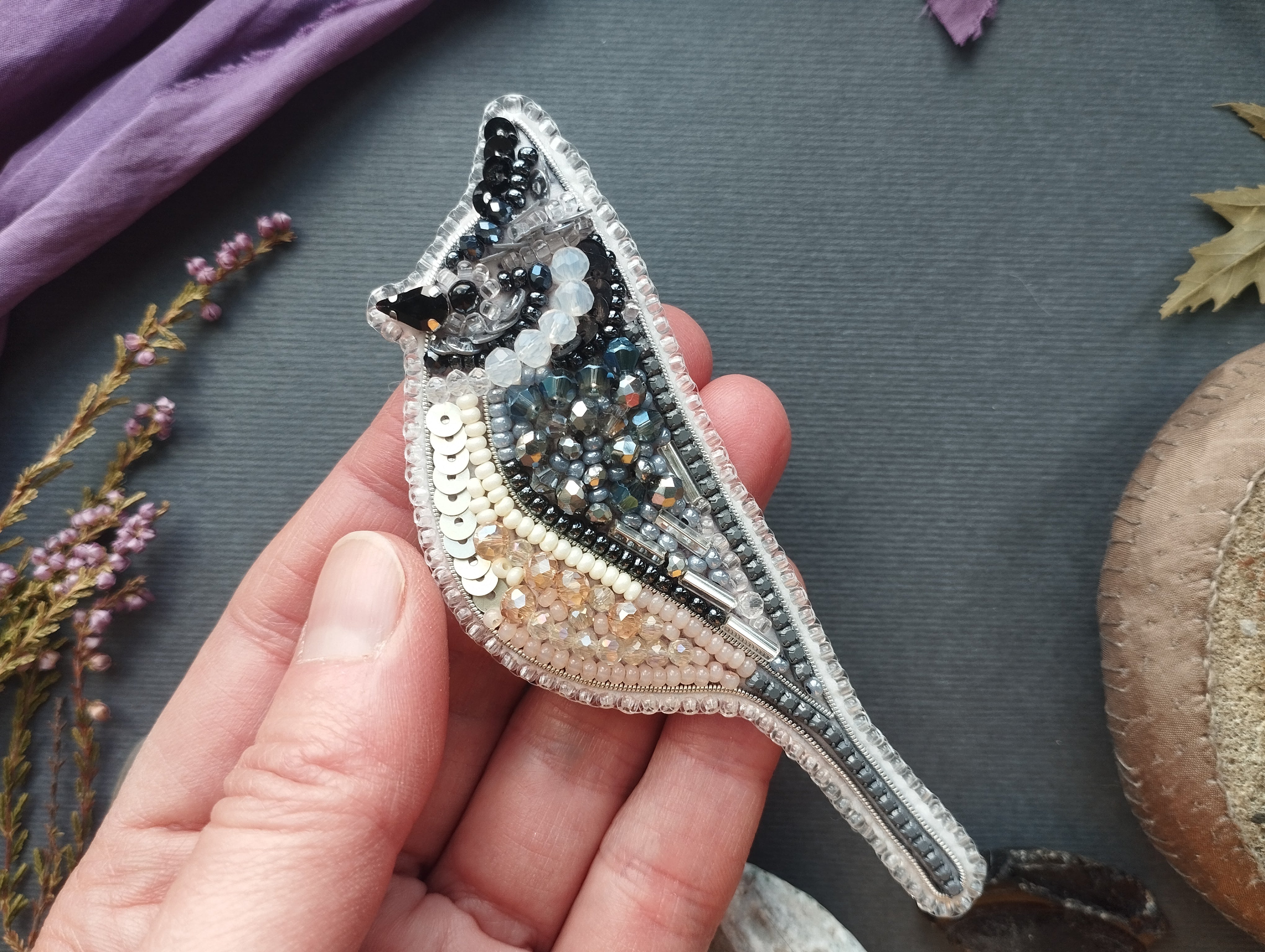 A hand is holding a decorative beaded bird brooch against a dark background.