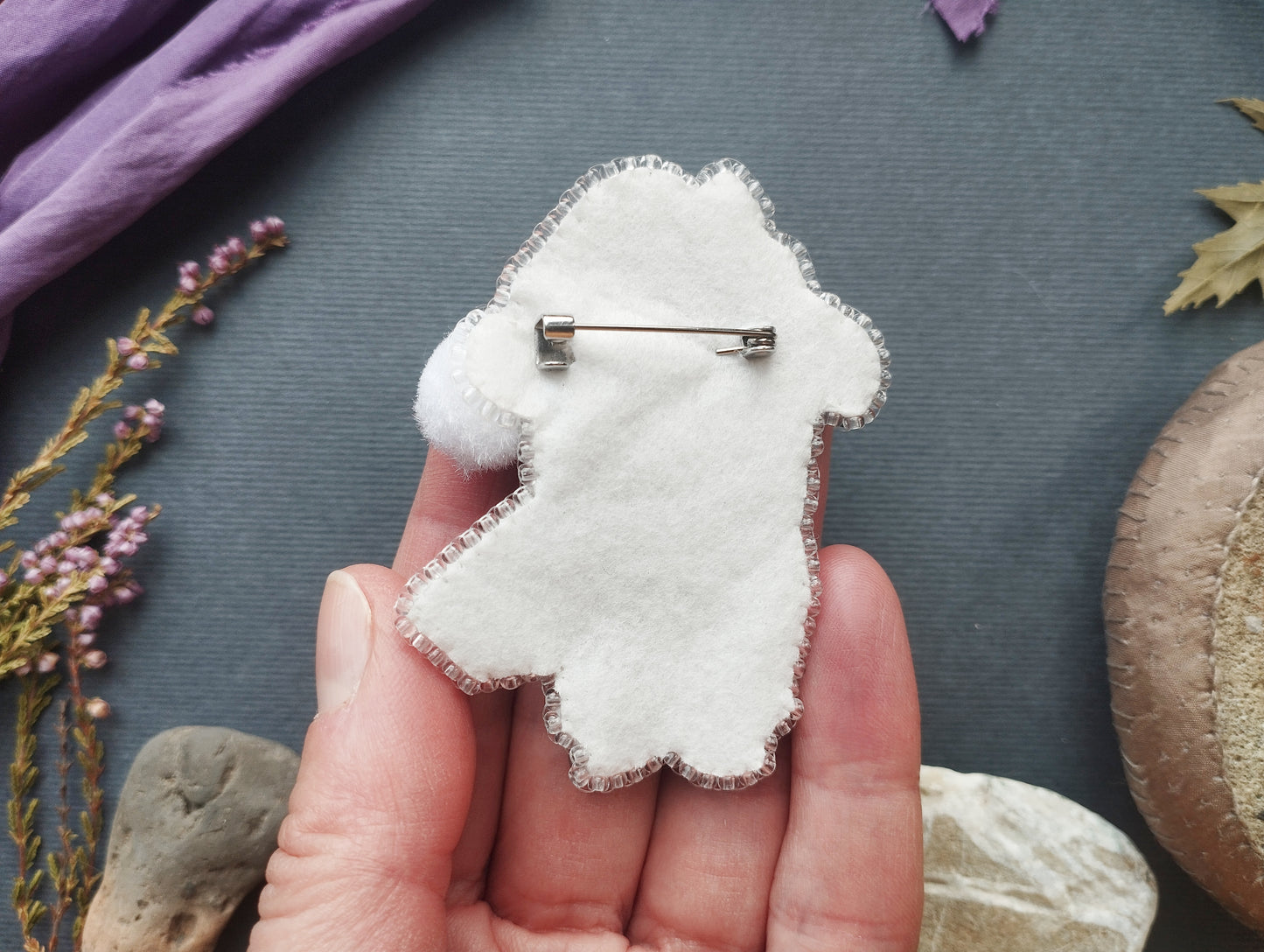 A hand holding a white felt animal pin against a dark background with various natural elements, such as flowers and rocks.