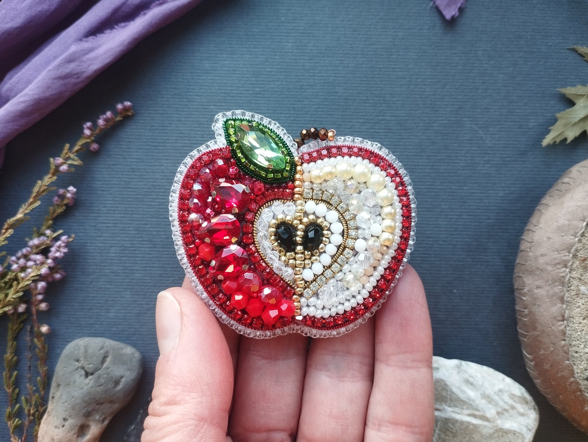 A hand holding a decorative apple-shaped brooch made of colorful beads and gemstones against a dark background.