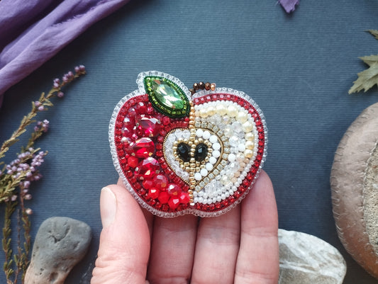 A hand holding a decorative apple-shaped brooch made of colorful beads and gemstones against a dark background.