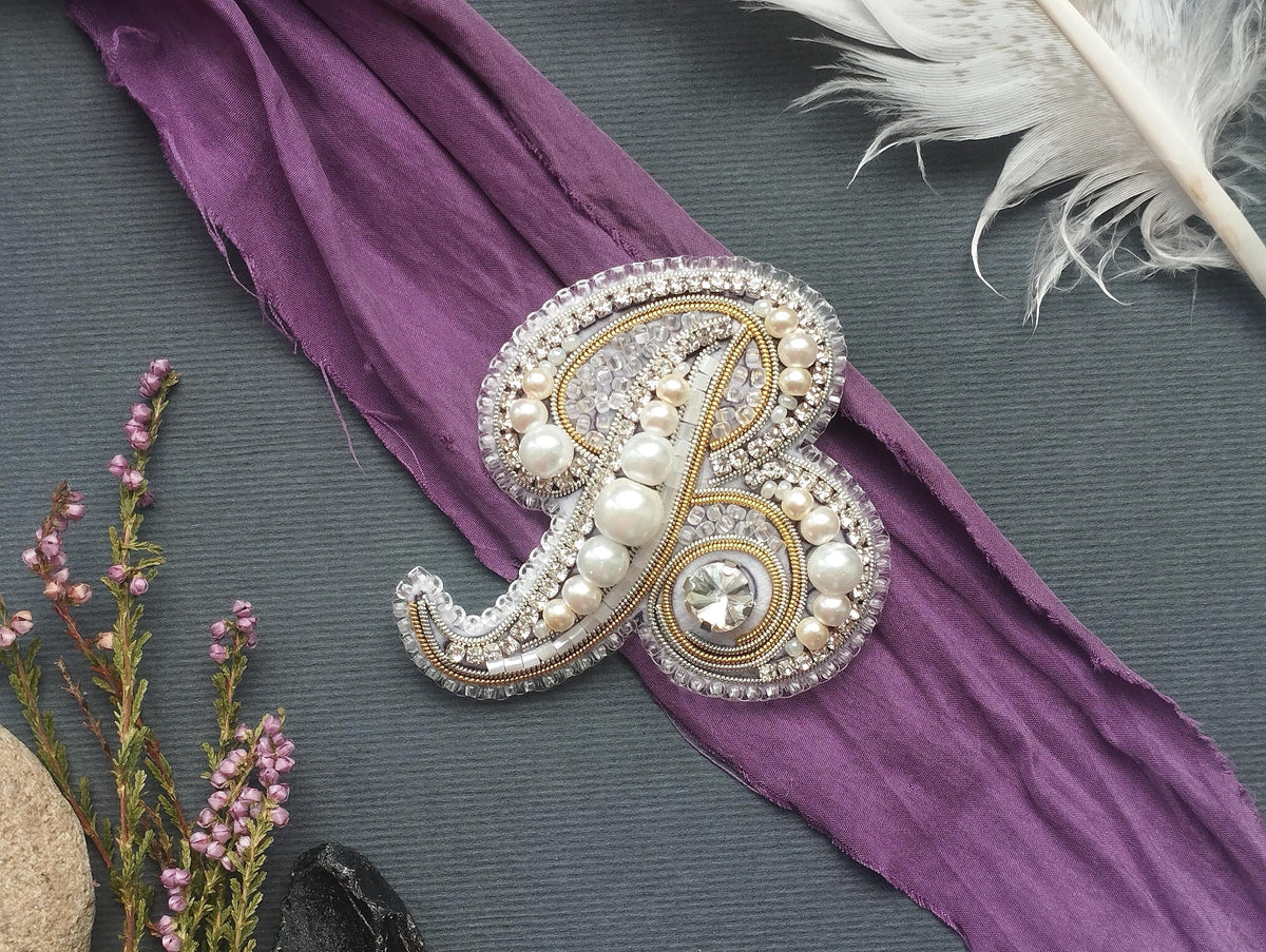 a decorative monogrammed letter "B" adorned with pearls and rhinestones, placed on a purple fabric.