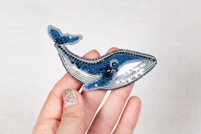 A hand holding a decorative blue and silver whale pin against a plain background.