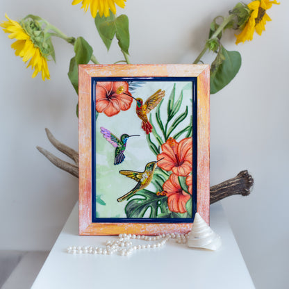 a framed painting of colorful hummingbirds surrounded by flowers, with a branch of sunflowers and a seashell on a white surface.