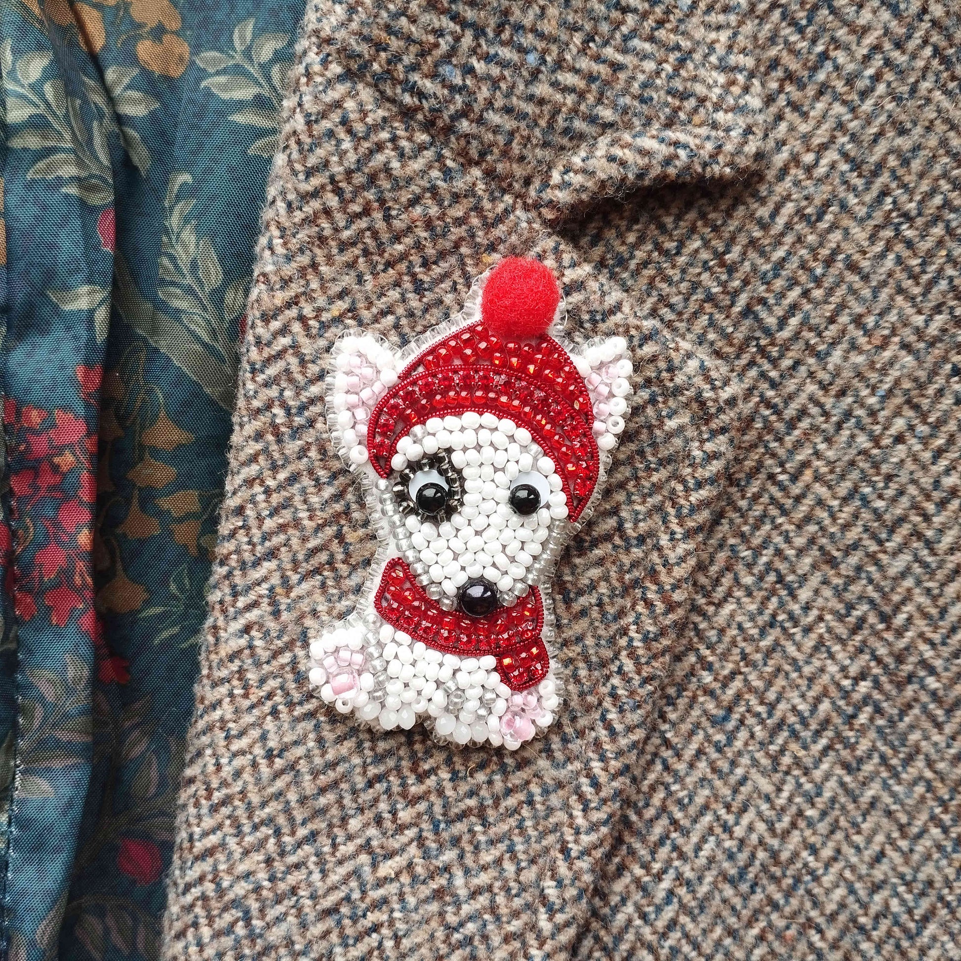 A close-up of a small, white dog-shaped brooch with a red hat and scarf, attached to a tweed jacket.
