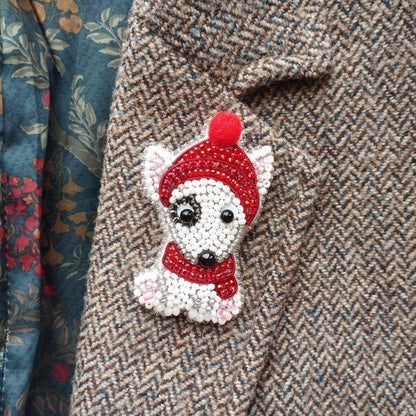 A close-up of a small, white dog-shaped brooch with a red hat and scarf, attached to a tweed jacket.