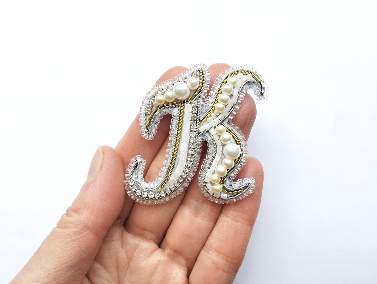 A hand holding a decorative letter 'K' made of beads, pearls, and rhinestones.