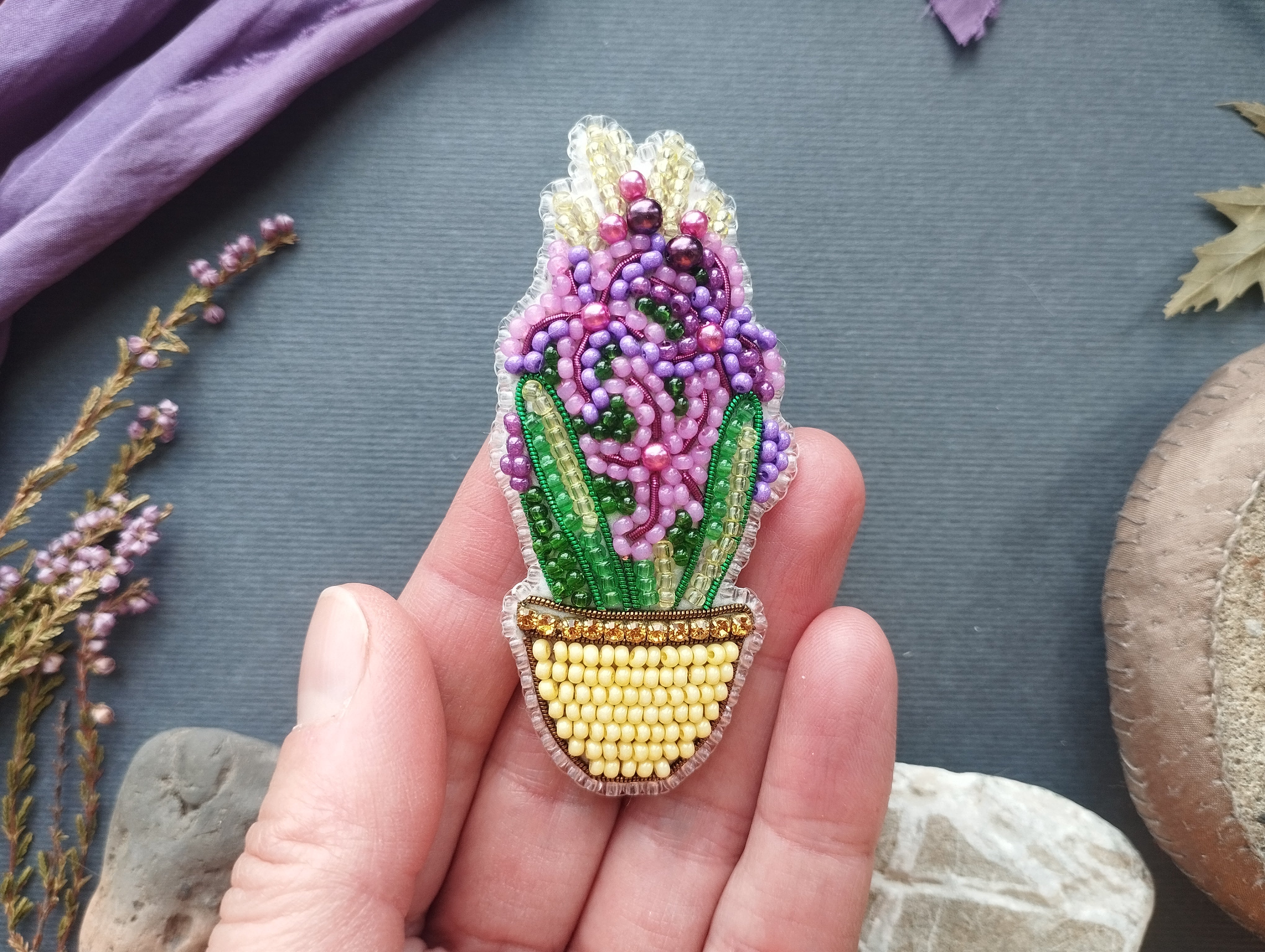 A hand holding a beaded embroidered piece depicting a flower in a pot, surrounded by various natural elements such as leaves and flowers.