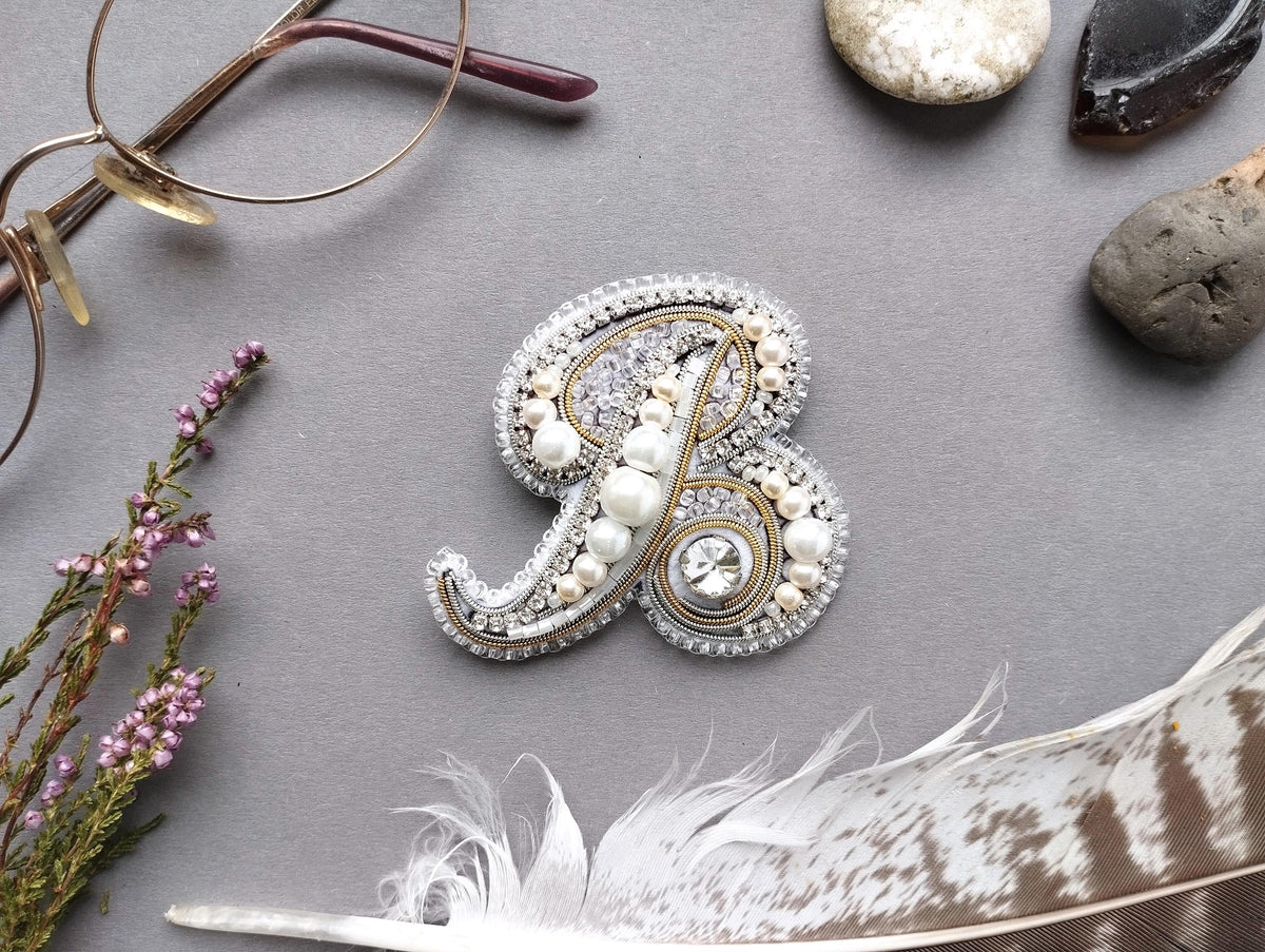 a close-up view of a decorative monogrammed letter "B" adorned with pearls and gemstones, surrounded by various accessories such as glasses, feathers, and rocks.