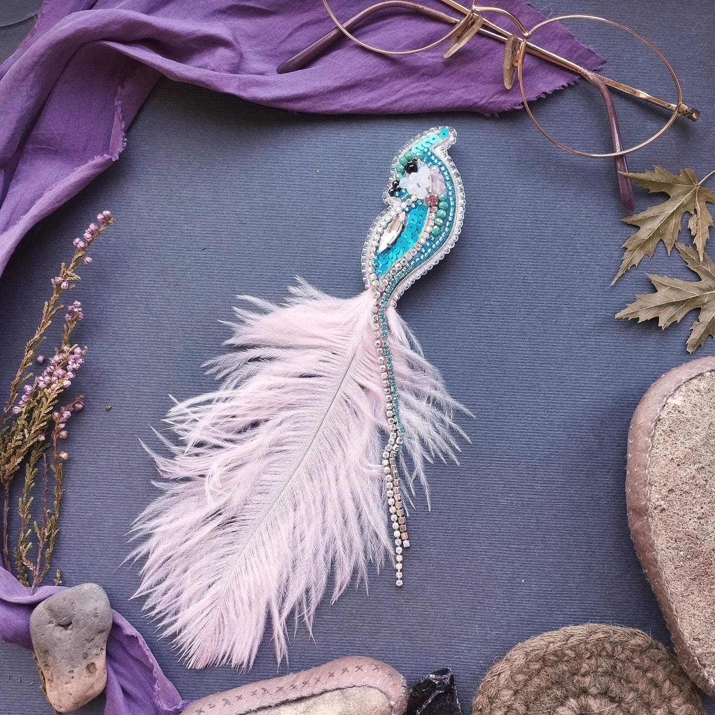 a close-up view of a feather with a beaded design, surrounded by various objects such as a pair of glasses, a purple cloth, and some rocks.