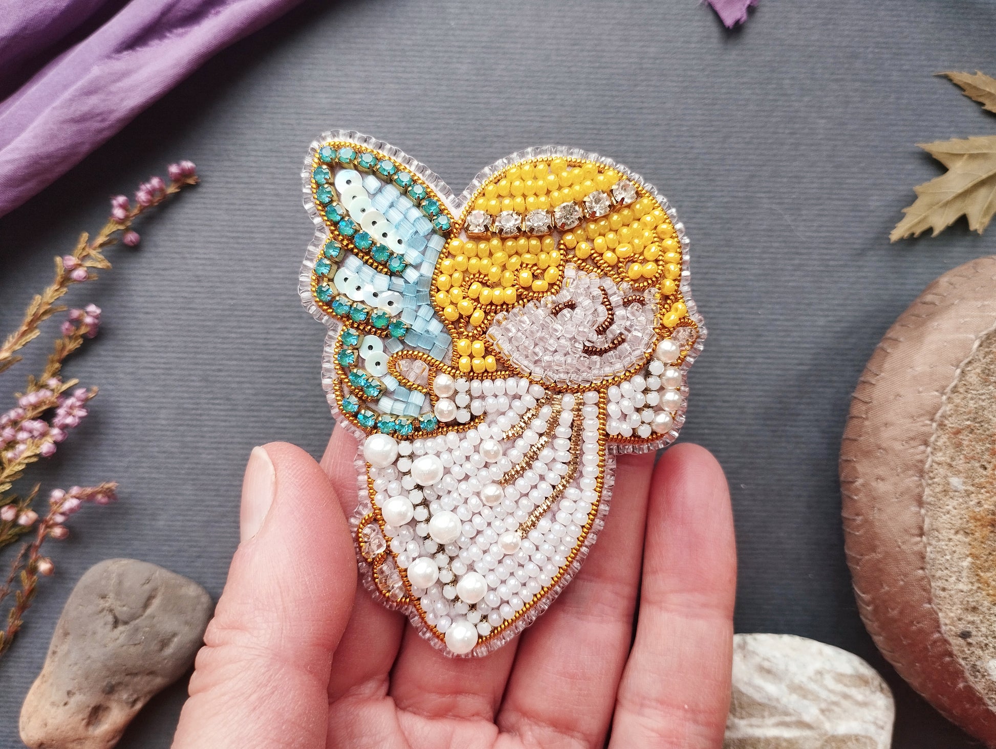 A hand holding a beaded embroidered figure of a smiling angel against a gray background.