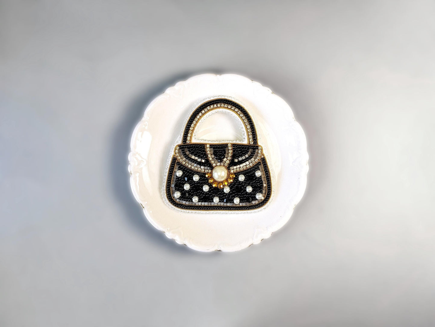 A decorative black and white handbag with a gold clasp and embellishments is displayed on a white plate against a gray background.