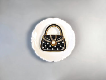 A decorative black and white handbag with a gold clasp and embellishments is displayed on a white plate against a gray background.