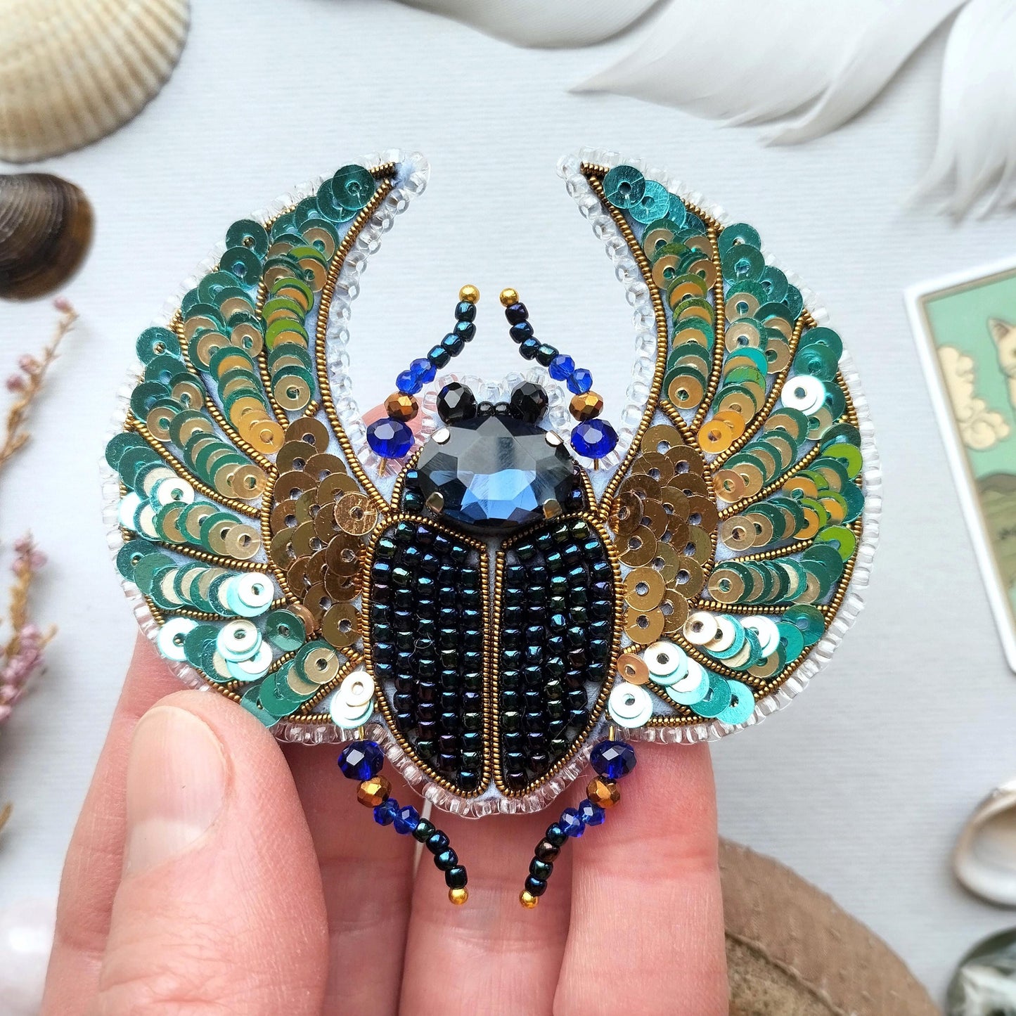 A hand is holding a decorative piece of jewelry featuring a detailed design of a large, colorful insect, possibly a beetle or a fly, with intricate patterns and sequins.