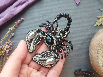 A hand holding a decorative black and silver beaded brooch with a scorpion design, surrounded by various natural elements such as flowers and leaves.