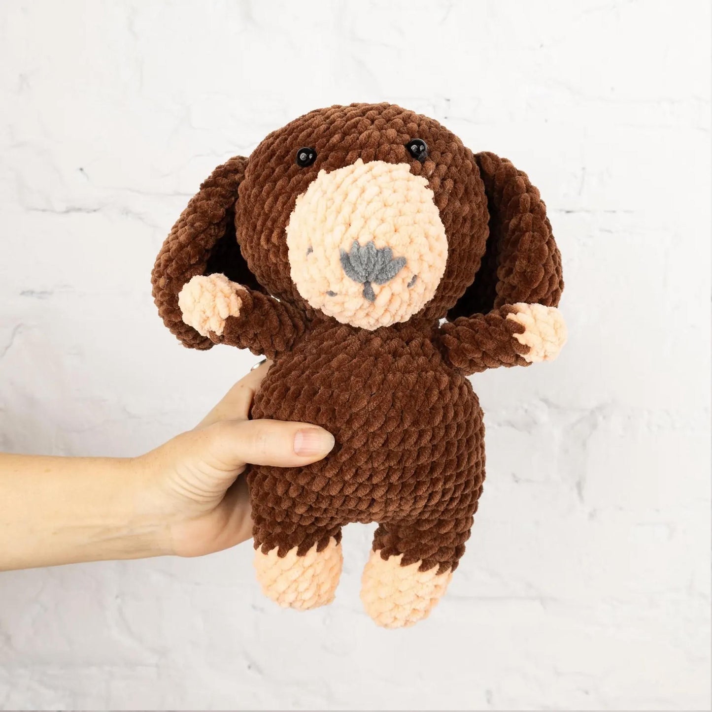 A hand is holding a brown, crocheted stuffed dog toy against a white brick wall.