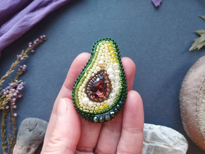 A hand holding a beaded avocado-shaped brooch with a gemstone in the center, surrounded by green and yellow beads.