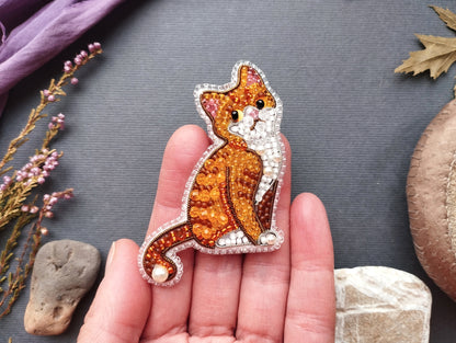 A hand holding a decorative cat pin made of beads and pearls against a gray background.