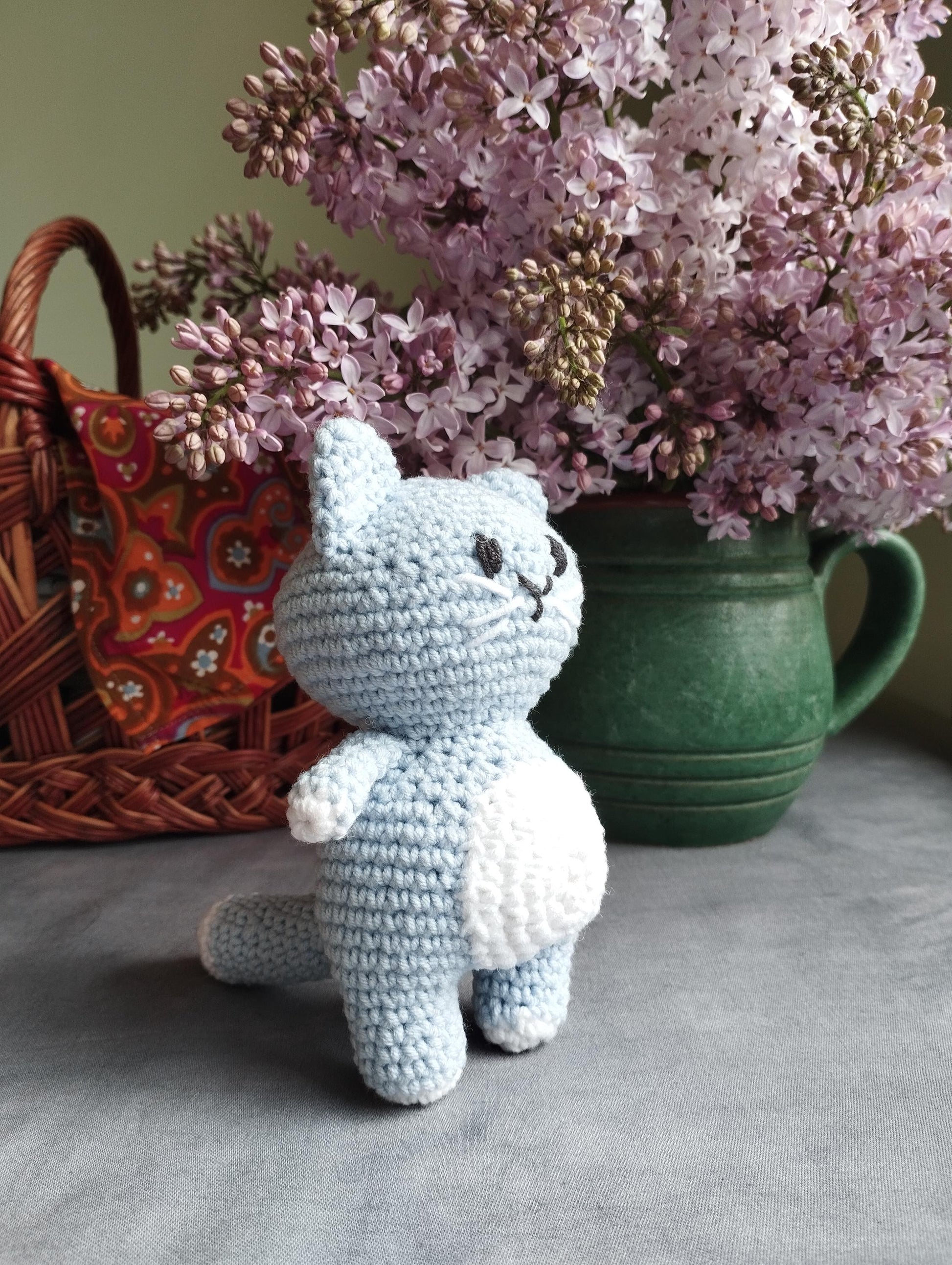 a crocheted cat sitting next to a basket of lilacs