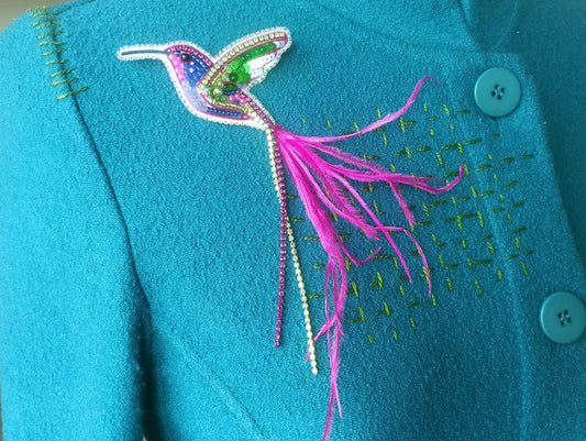 A close-up image of a teal-colored jacket with a decorative bird brooch featuring a colorful hummingbird design and a long pink feather.