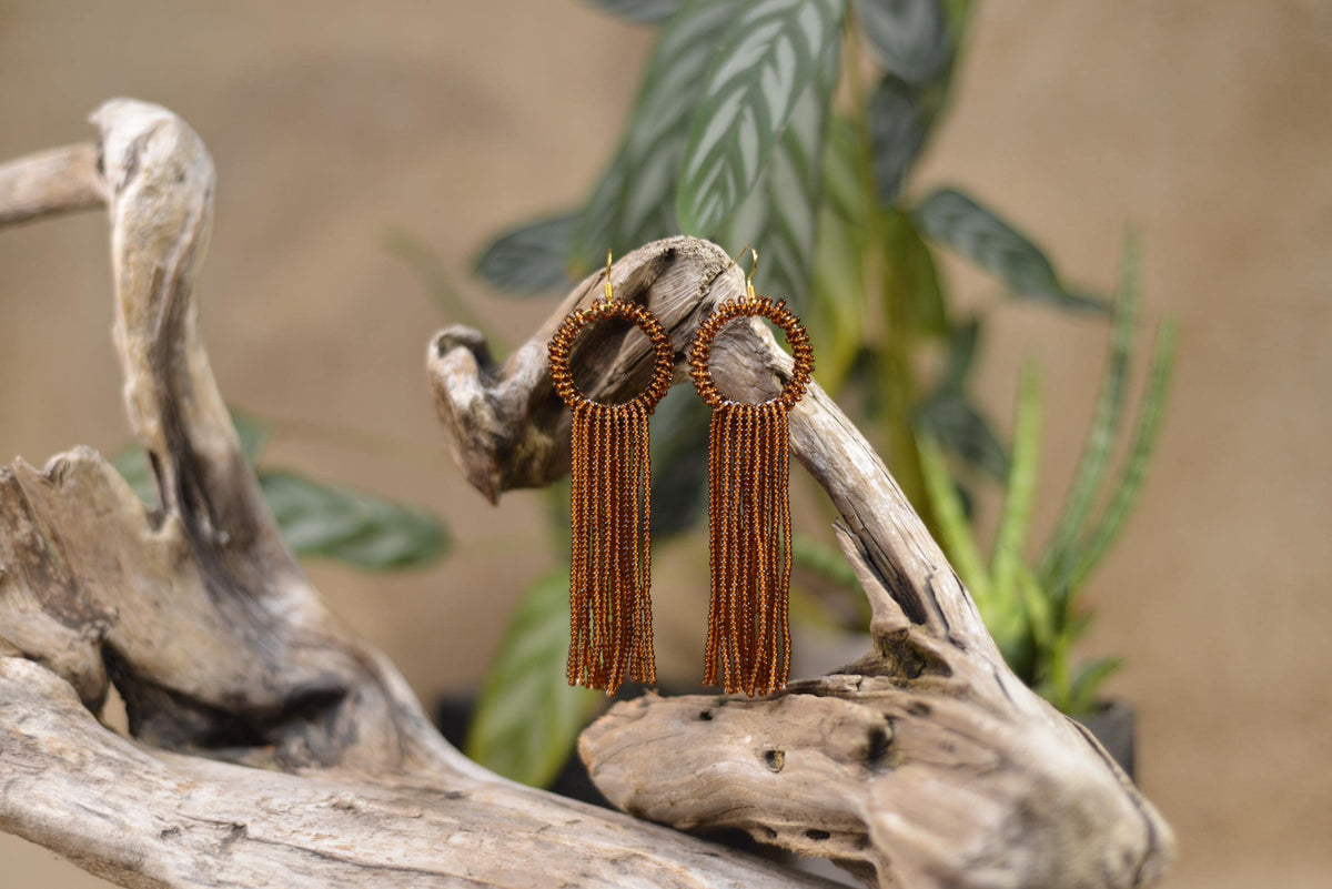 A pair of brown beaded earrings with long tassels hanging from them, displayed on a wooden branch or log against a blurred background of plants.