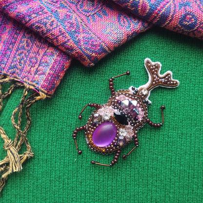 a colorful, intricately designed insect-shaped brooch with purple gemstones and gold accents, placed on a green textured surface.