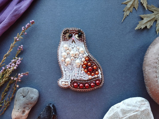 a decorative cat-shaped brooch made of beads and pearls, surrounded by various natural elements such as leaves, rocks, and flowers on a blue background.