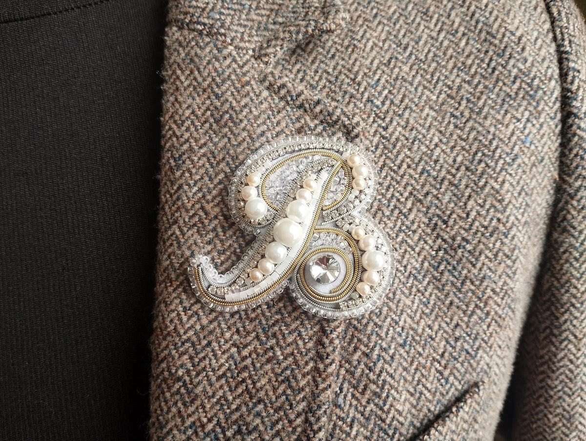A close-up image of a decorative monogram brooch on a tweed jacket, featuring a large letter "B" with pearls and a crystal accent.