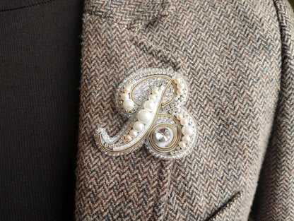 A close-up image of a decorative monogram brooch on a tweed jacket, featuring a large letter "B" with pearls and a crystal accent.