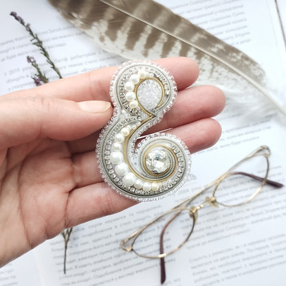 A hand holding a decorative brooch with pearls and a crystal, surrounded by a feather and a pair of glasses on a page of text.
