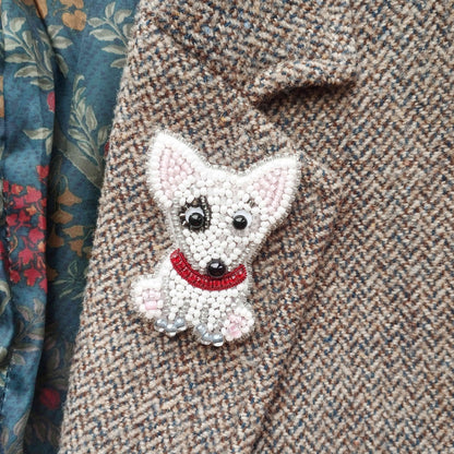 A close-up image of a small, white dog-shaped embroidered piece on a tweed fabric, with a red collar.