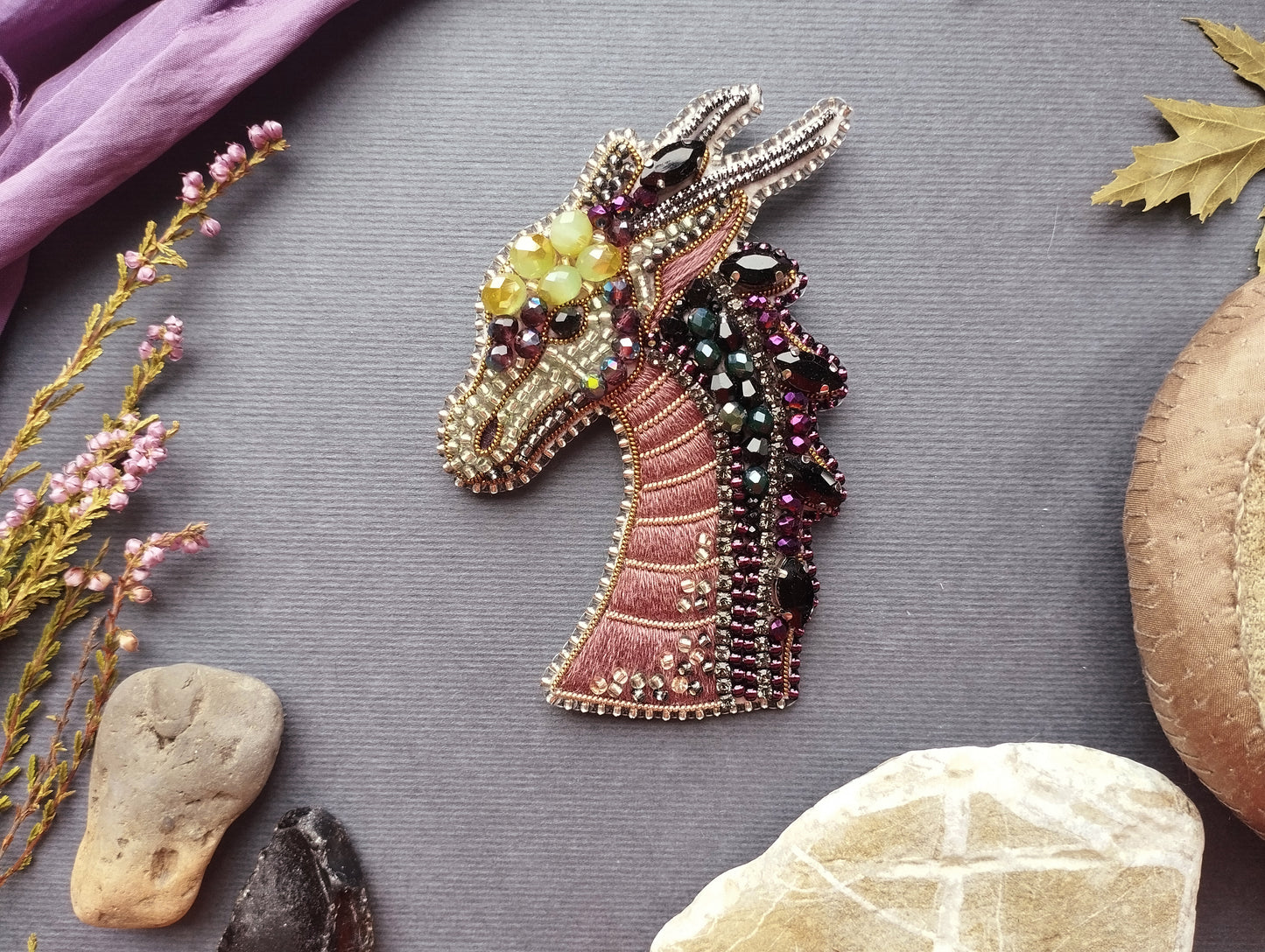 a decorative dragon brooch with intricate beading and gemstone embellishments, surrounded by various natural elements such as leaves, rocks, and flowers.