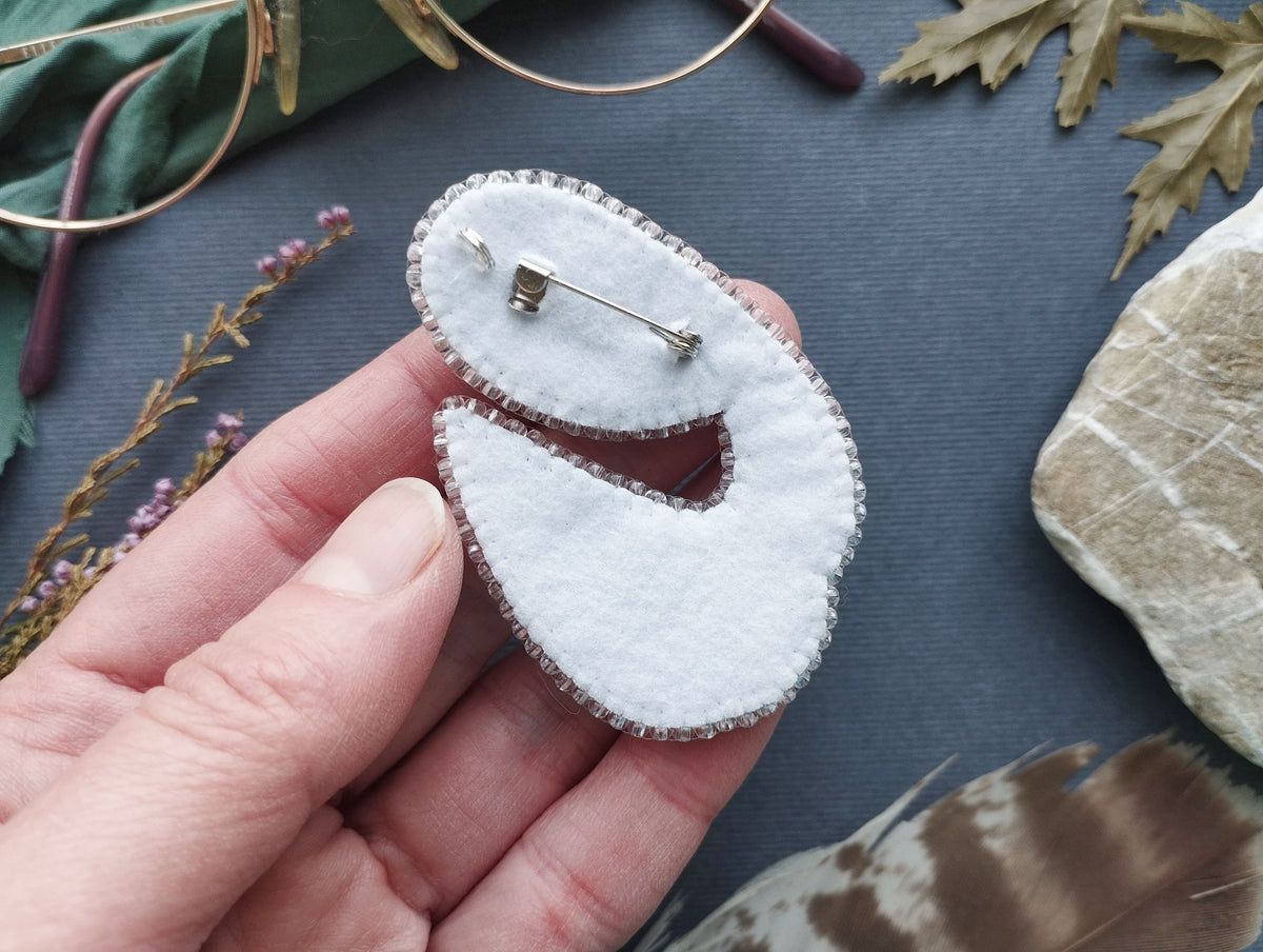 A hand holding a white felt pin with a silver pin attached to it, surrounded by various objects such as a pair of glasses, leaves, and feathers.