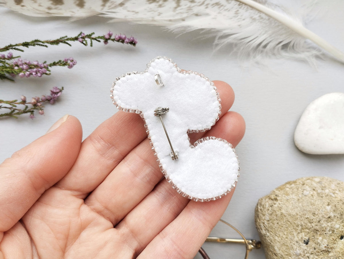 A hand holding a white felt object with a silver pin attached to it, surrounded by various natural elements such as feathers, flowers, and rocks.
