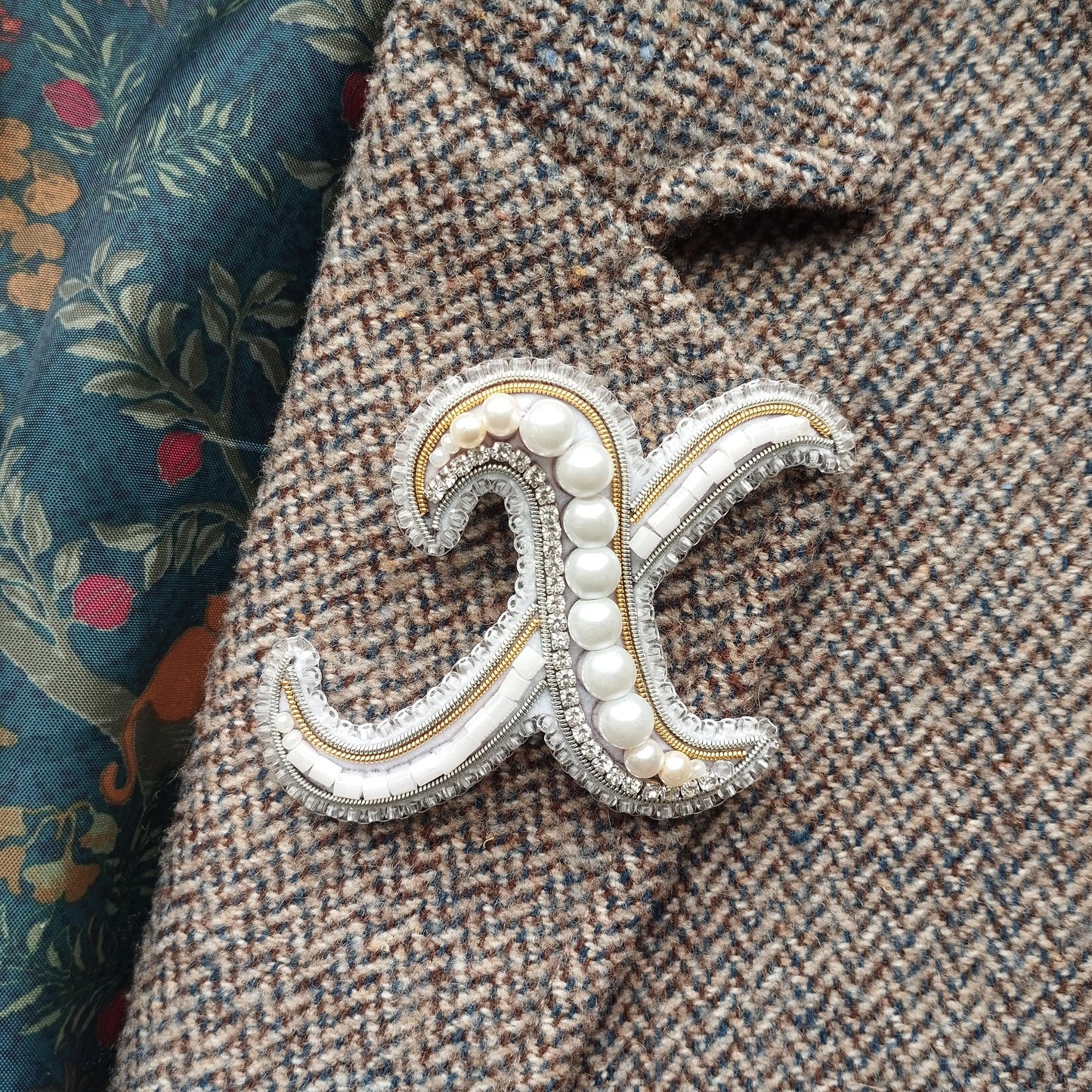 A close-up image of a decorative monogram brooch with pearls and a metallic sheen, placed on a tweed jacket.