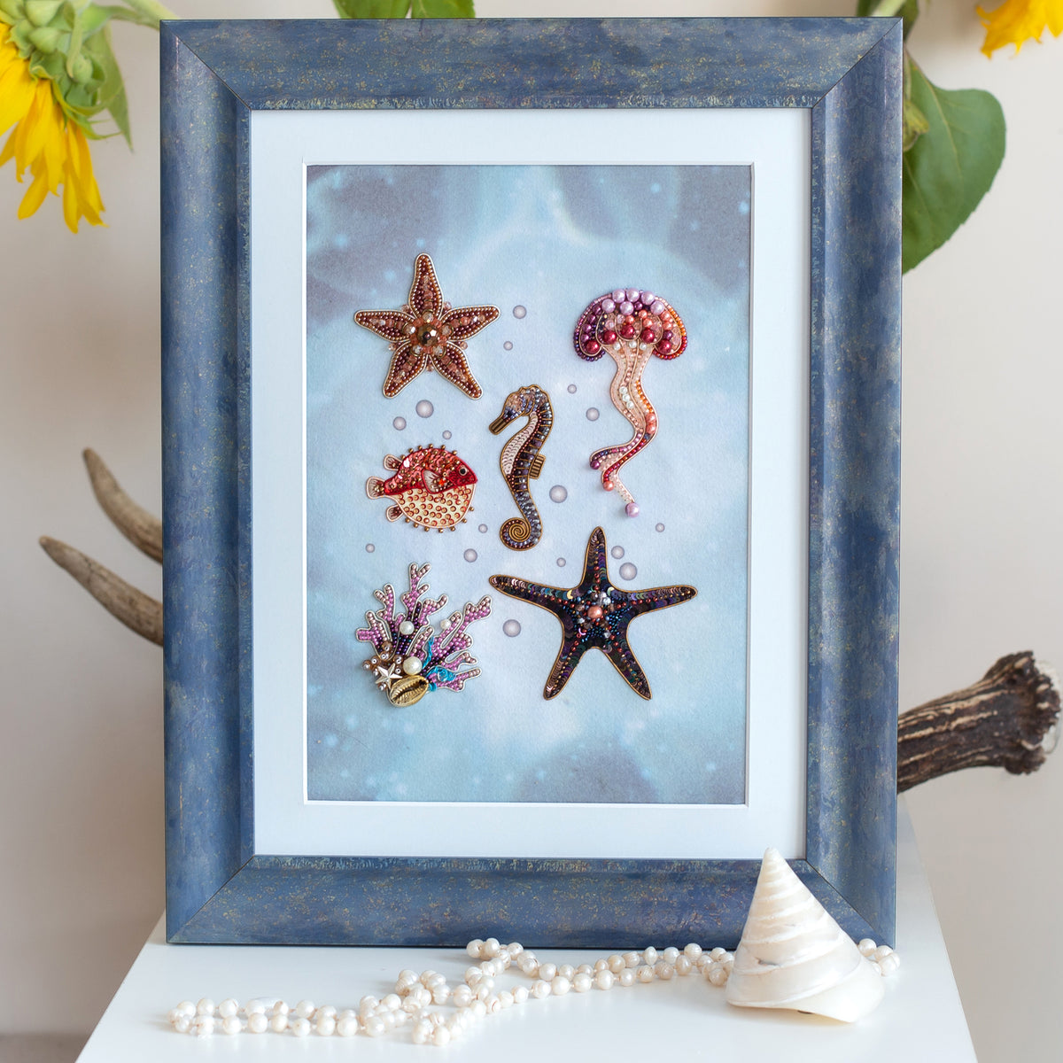 a framed collage of various sea creatures, including a starfish, a jellyfish, a seahorse, and a pearl. The frame is blue and the background is a light blue color. The image also includes a small white shell and some pearls.