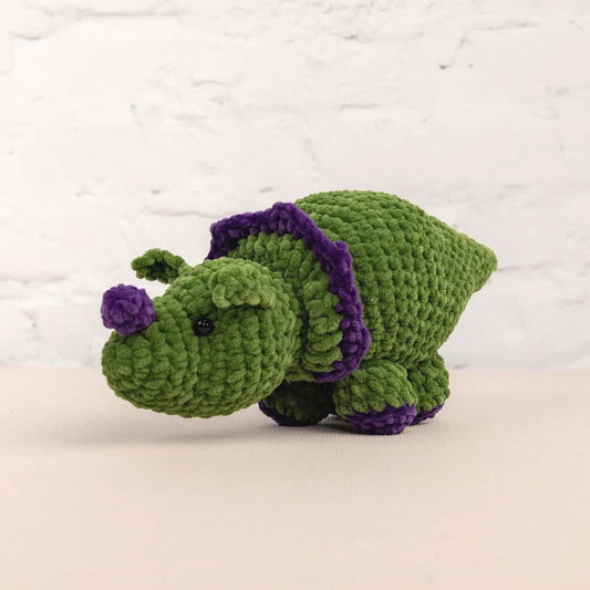 A green crocheted toy resembling a rhinoceros with purple accents is sitting on a white surface against a white background.