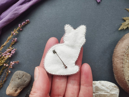 A hand holding a white felt cat pin against a dark background with various objects, including a purple fabric, flowers, and rocks.