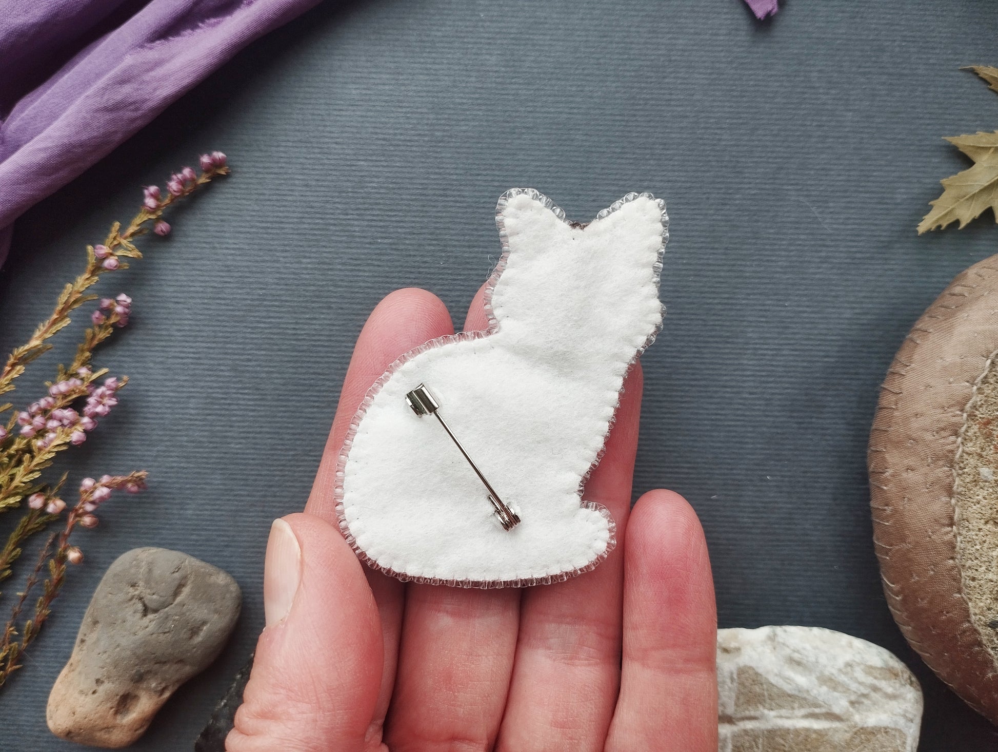 A hand holding a white felt cat pin against a dark background with various objects, including a purple fabric, flowers, and rocks.
