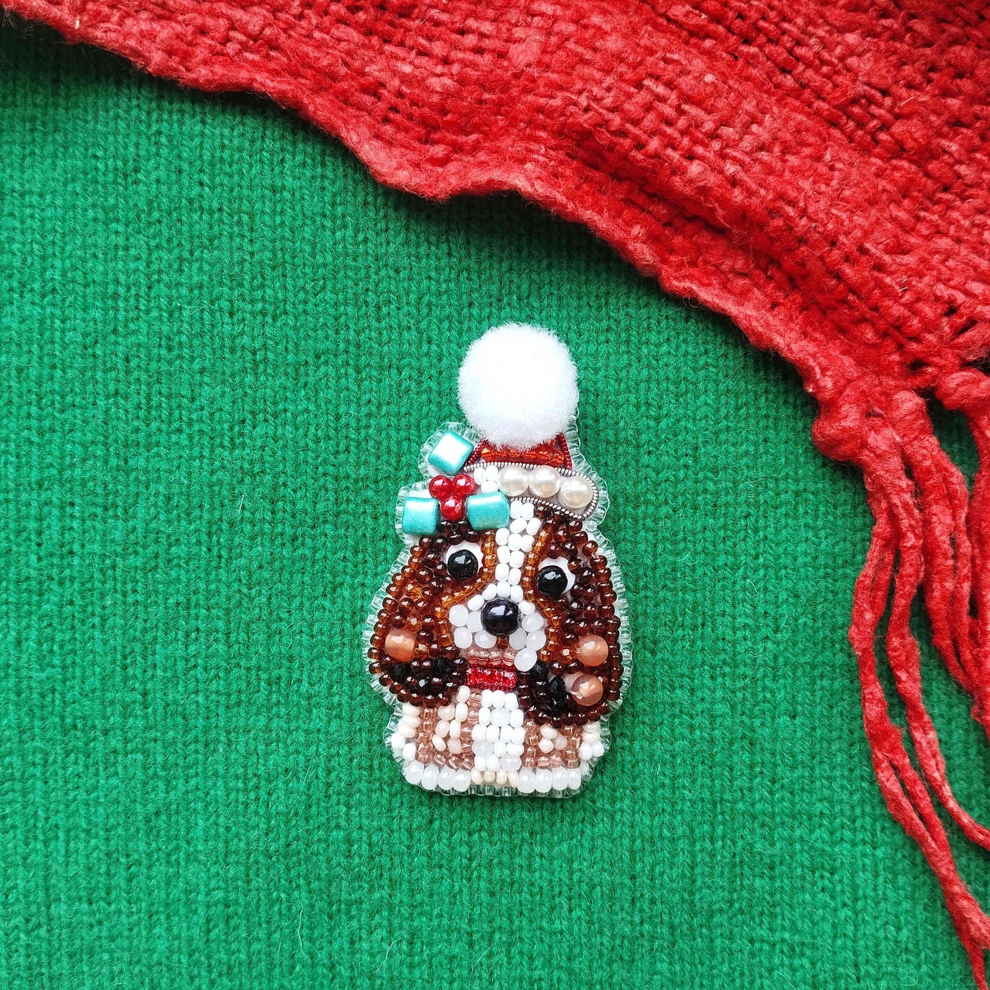 A small, intricately beaded dog brooch is displayed against a green background, with a red scarf visible on the right side.