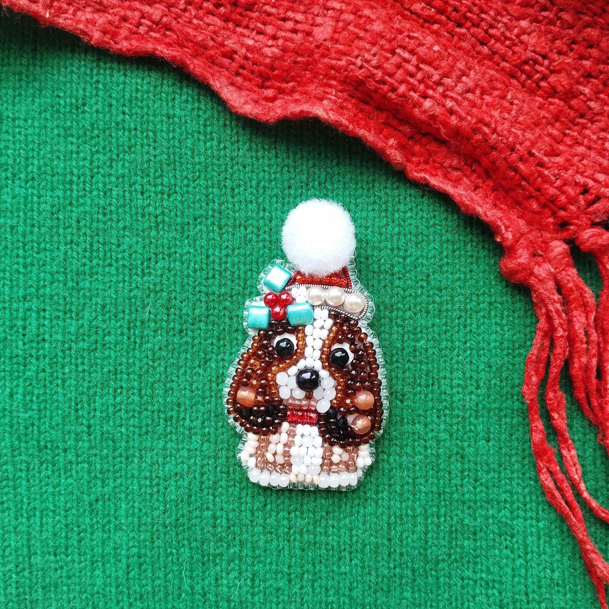 A small, intricately beaded dog brooch is displayed against a green background, with a red scarf visible on the right side.