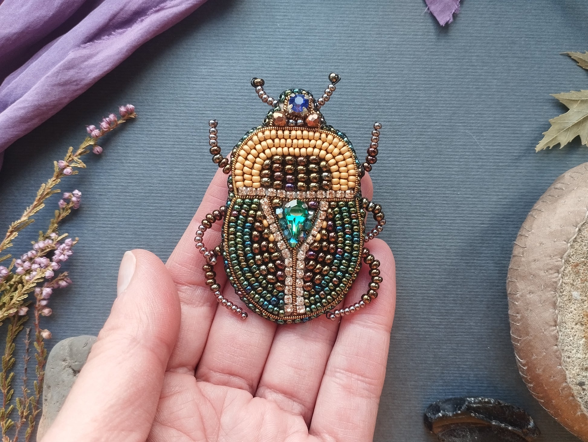 A hand holding a beaded, embroidered, and bejeweled insect-shaped brooch against a blue background with various natural elements, such as flowers and leaves.