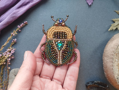 A hand holding a beaded, embroidered, and bejeweled insect-shaped brooch against a blue background with various natural elements, such as flowers and leaves.