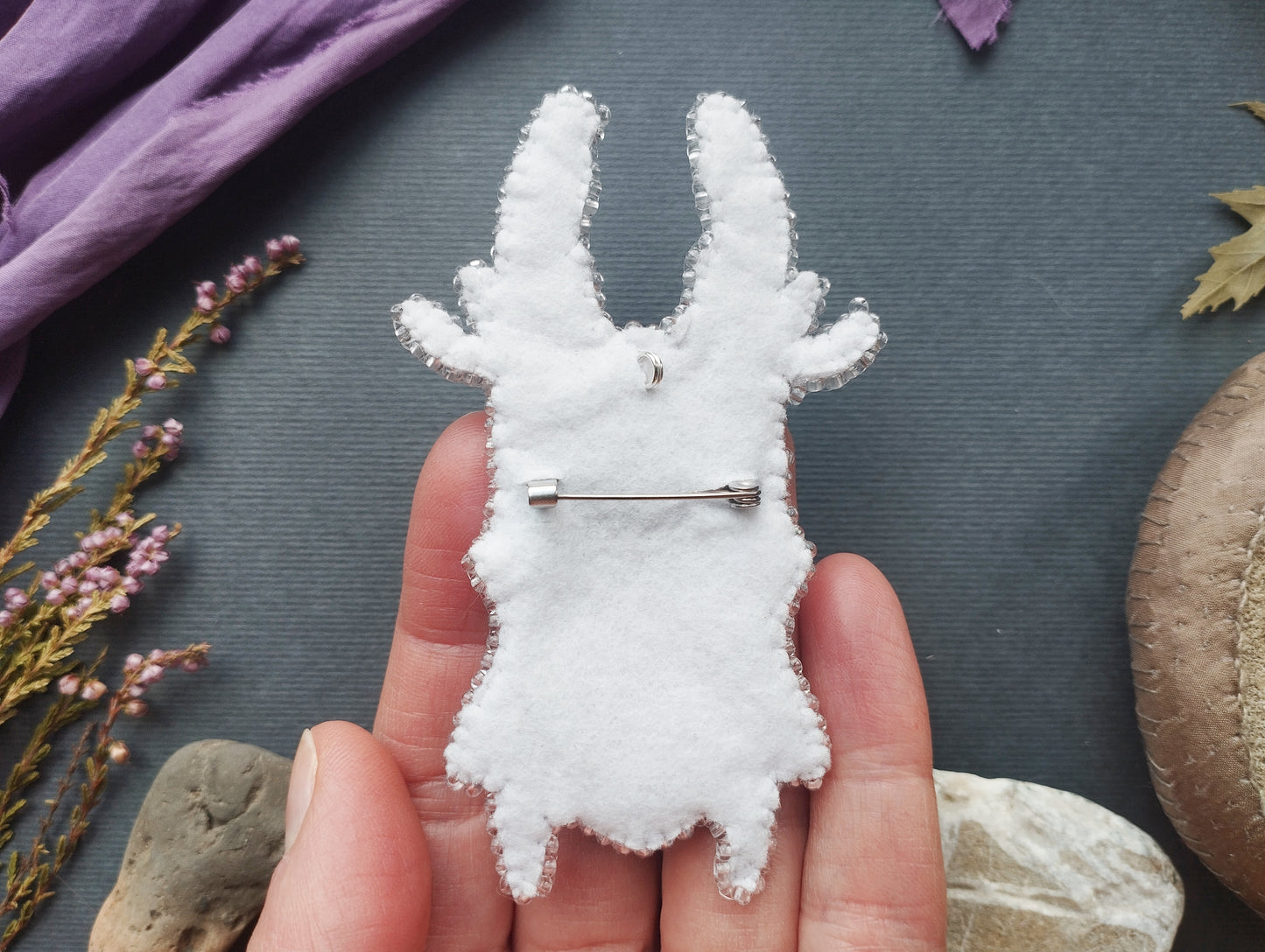 A hand holding a white fabric object with a pin on it, surrounded by various objects such as flowers, rocks, and a purple fabric.