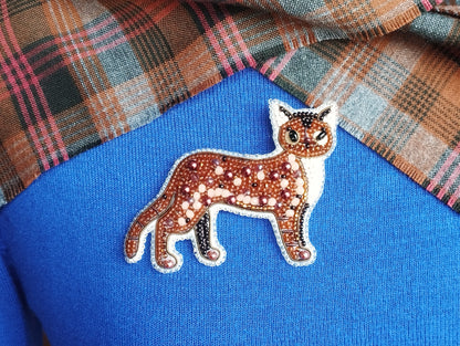 A close-up image of a beaded cat pin on a blue fabric background.