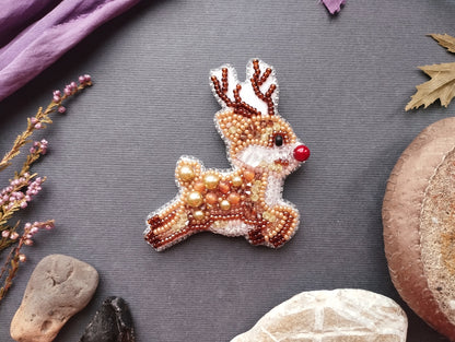 a beaded reindeer brooch with a red nose, placed on a gray background among various objects such as leaves, rocks, and a mushroom.