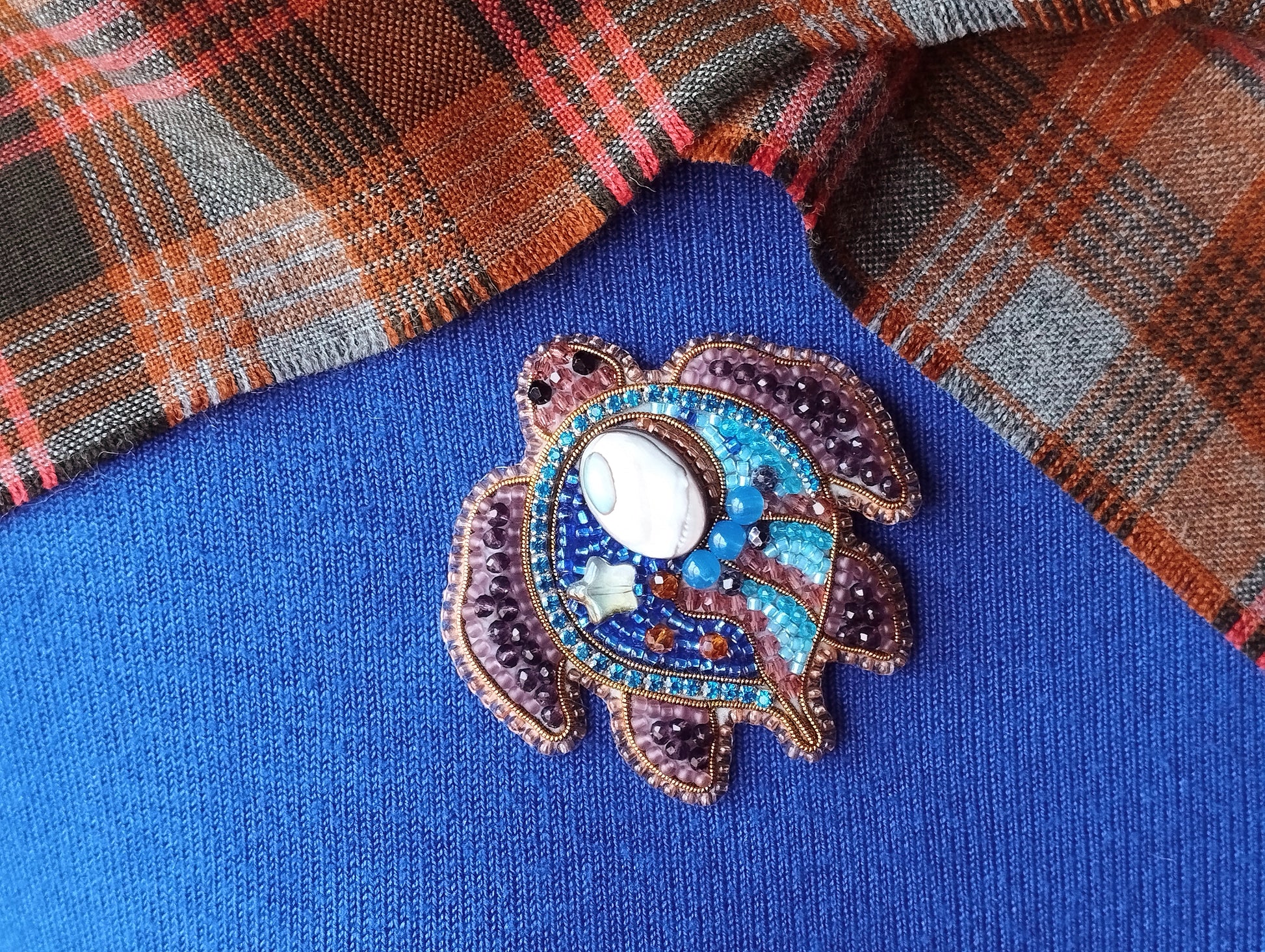 A decorative turtle brooch with a white shell and colorful beads is displayed on a blue fabric background.